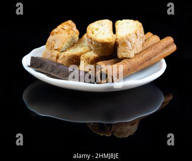 Cinnanon bastoni, cioccolato e bisquits in una piastra bianca su sfondo nero riflettente Foto Stock