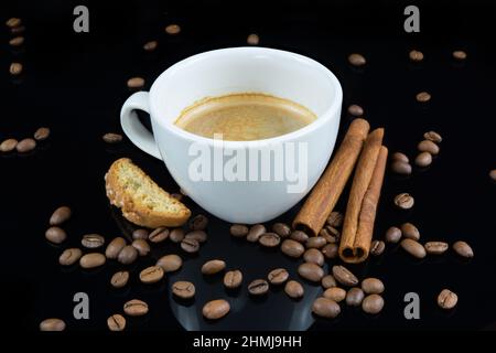 Un caffè in tazza bianca circondato da chicchi di caffè e bastoncini di cannella su sfondo nero riflettente Foto Stock