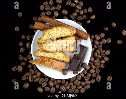 Bastoncini Cinnanon, cioccolato e bisquits in un piatto bianco circondato da chicchi di caffè su sfondo nero riflettente Foto Stock