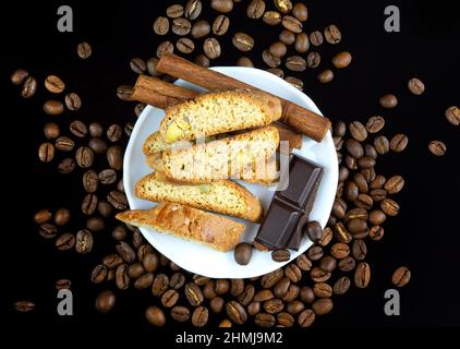 Bastoncini Cinnanon, cioccolato e bisquits in un piatto bianco circondato da chicchi di caffè su sfondo nero riflettente Foto Stock