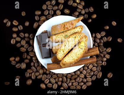 Bastoncini Cinnanon, cioccolato e bisquits in un piatto bianco circondato da chicchi di caffè su sfondo nero riflettente Foto Stock