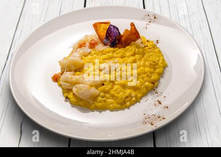 Risotto allo zafferano giallo con seppie e polpa di pomodoro fresco, ricetta gourmet in piatto bianco su tavola di legno bianco Foto Stock