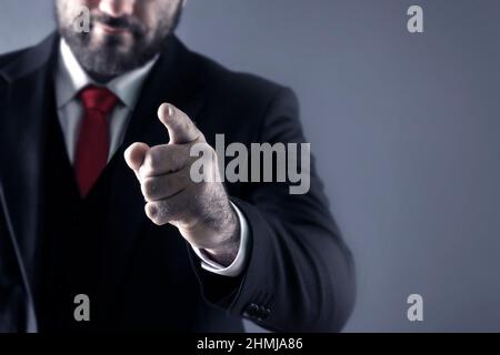 Dito sulla fotocamera. Gesto di puntamento. Tu. Un uomo in vestito punta il dito in avanti su uno sfondo grigio. Foto Stock