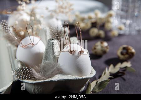 Concetto di Pasqua. Un uovo bianco con orecchie di filo di rame e un viso di coniglio dipinto in una scatola. Partridge uova e piume. Foto Stock