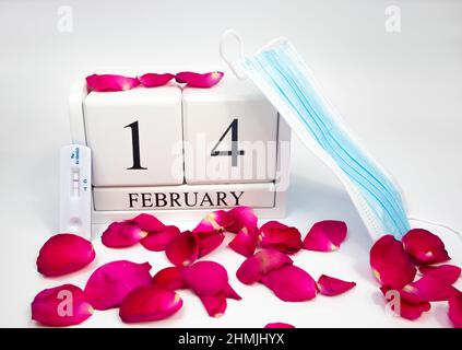 Febbraio 14 calendario in legno bianco su sfondo bianco con petali di rosa rossa. Accanto a lui c'è una maschera medica e un test positivo covid Foto Stock