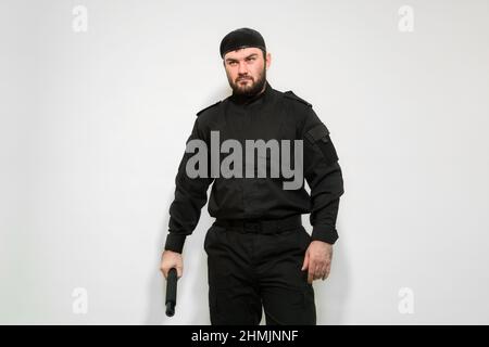 Guardia di sicurezza uomo in uniforme nera con un batone nelle sue mani su uno sfondo bianco. Mercenario. Sicurezza privata. Bodyguard. Difensore. Foto Stock