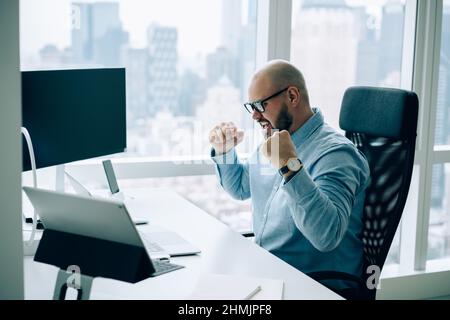 Felice mazziere maschio che alza i pugni in soddisfazione in ufficio moderno Foto Stock