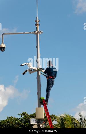 Un lavoratore maschio su una scala metallica installa telecamere di sicurezza vicino a una spiaggia pubblica in Messico Foto Stock