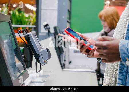 Uomo nero in giacca calda in denim utilizza lo smartphone per pagare l'acquisto presso il punto self-check in vista supermercato Foto Stock