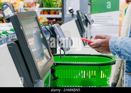 Uomo nero in giacca calda in denim utilizza lo smartphone per pagare l'acquisto presso il punto self-check in vista supermercato Foto Stock