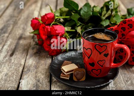 Bouquet di rose rosse, tazza di caffè, scatola regalo e caramelle al cioccolato su tavola di legno. Concetto romantico per San Valentino. Foto Stock