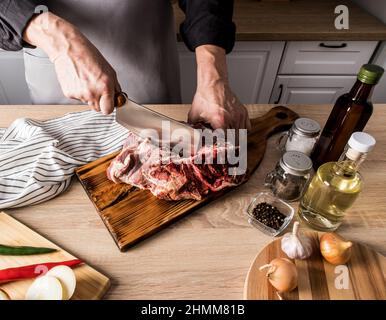 un'immagine senza volto di un uomo in un grembiule grigio con un'ascia taglia un grosso pezzo di manzo sulle ossa. il concetto di cottura della carne in cucina a casa Foto Stock