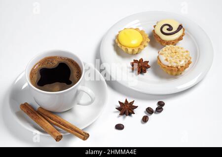 Tazza di caffè, piccoli muffin smaltati, bastoncini di cannella e stella di anice su sfondo bianco Foto Stock