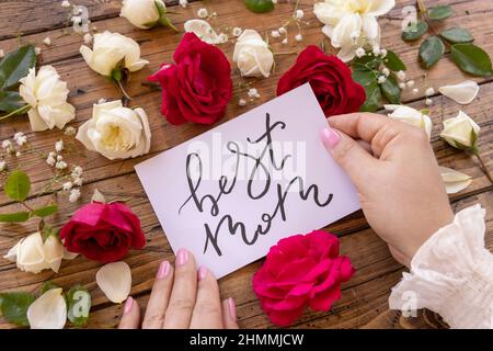 Mani con carta manoscritta MIGLIOR MOM circondato da rose rosse e crema da vicino su un tavolo di legno. Femminine romantica dichiarazione di amore vicino ai fiori Foto Stock