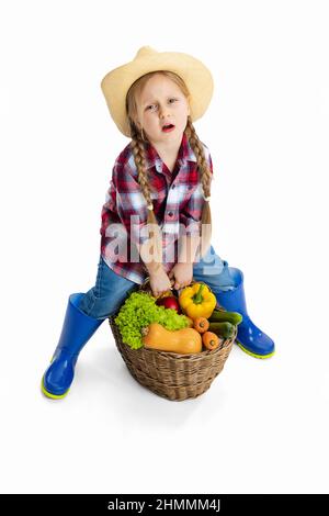 Scatto dello studio della ragazza piccola carina, capretto emotivo nell'immagine del coltivatore, giardiniere che tiene il cestino grande delle verdure isolato su sfondo bianco. Modello in Foto Stock