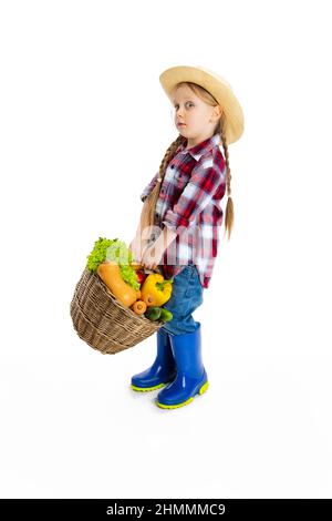 Scatto dello studio della ragazza piccola carina, capretto emotivo nell'immagine del coltivatore, giardiniere che tiene il cestino grande delle verdure isolato su sfondo bianco. Modello in Foto Stock