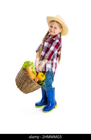 Scatto dello studio della ragazza piccola carina, capretto emotivo nell'immagine del coltivatore, giardiniere che tiene il cestino grande delle verdure isolato su sfondo bianco. Modello in Foto Stock