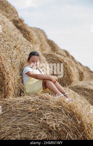 Ritratto di piccola ragazza Sly che riposa su haystack guardando la macchina fotografica con gli occhi squinted che indossa la Sundress con dozzine di haystack sullo sfondo. Lontano da Foto Stock