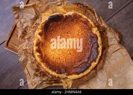 Vista dall'alto di una vita morta di una cheesecake fresca dal forno su carta marrone e asse di legno. Concetto di gastronomia spagnola tradizionale. Foto Stock