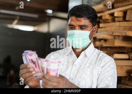 Metta a fuoco su mano, carpentiere professionista indiano giovane con maschera medica che conta i soldi al negozio - concetto di profitto, legno o guadagni del lavoratore dell'artigianato Foto Stock