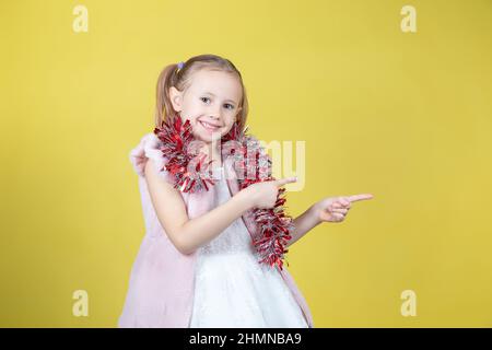 Ritratto di bambina carina in un abito elegante e con tinsel facendo un gesto di puntamento con posto per il testo su sfondo giallo. Il concetto Foto Stock