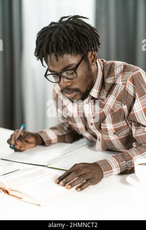 Lo studente afro-americano legge il libro e prende appunti nel suo copybook facendo i suoi compiti. Ritratto giovane uomo in occhiali studio a casa o in biblioteca. Concetto educativo. Foto Stock