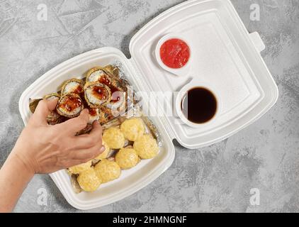 Deliziosi panini di sushi in scatole di plastica usa e getta. La mano prende un rotolo cotto con bacchette. Concetto di servizio di consegna degli alimenti, impilamento piatto Foto Stock