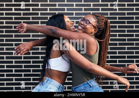 coppia di giovani ragazze nere sorridenti felice abbracciandosi davanti ad un muro di mattoni, concetto di gioventù e amicizia, spazio di copia per il testo Foto Stock