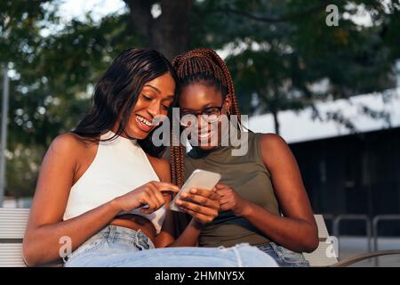due amici neri che si divertono seduti su una panca del parco guardando un telefono cellulare, concetto di gioventù e comunicazione Foto Stock