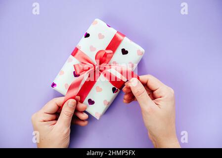 Le mani che slegano un arco a nastro su una scatola regalo avvolta in carta di disegno di cuore Foto Stock