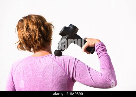 La donna applica un massaggio con una pistola a percussione sulle spalle, isolata su sfondo bianco. Foto Stock