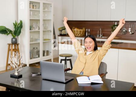 La giovane donna asiatica sorprende la vittoria e guarda il portatile seduto a casa. Femmina celebra le buone notizie ricevute via e-mail, ha ottenuto un'offerta di lavoro. Donna stupita gioire vincere in una lotteria online Foto Stock