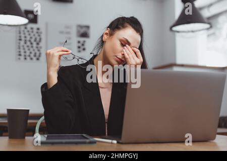 La giovane donna sente l'affaticamento degli occhi durante il lavoro sul laptop. Togliere gli occhiali e massaggiare gli occhi Foto Stock