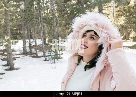 Donna sorridente che indossa un cappotto invernale con cappa in pelliccia sintetica in piedi in una foresta innevata, Spagna Foto Stock