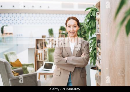 Donna business bruna sorridente per adulti quarant'anni con capelli lunghi in elegante abito beige e jeans al luogo pubblico, verde ufficio open space, coworking. Foto Stock