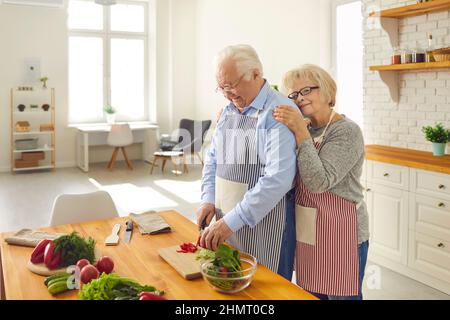 Felice amorevole coppia anziana in grembiuli che cucinano una sana cena vegetariana insieme e abbraccio Foto Stock