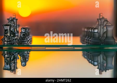 Scacchi di vetro posti su un tavolo di vetro visto dal lato con un riflesso orizzontale e sfondo arancione con un cerchio solare giallo tramonto. Foto Stock