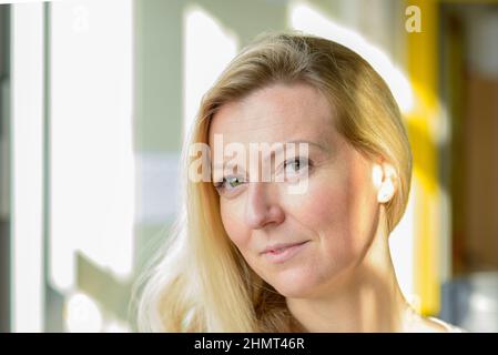 Donna bionda scettica che guarda sospettosamente alla fotocamera con un'espressione meditata vicino a una finestra con luce solare riflessa sul suo viso Foto Stock