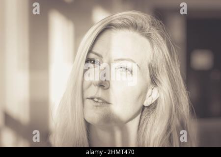 Donna bionda scettica che guarda sospettosamente alla fotocamera con un'espressione meditata vicino a una finestra in bianco e nero Foto Stock