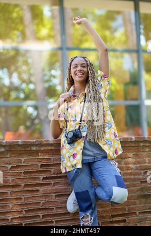 Donna nera con trecce africane, sollevando il braccio con gioia. Ragazza che tiene una macchina fotografica. Foto Stock