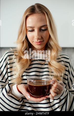 Ritratto di giovane ragazza felice bionda sorridente donna che tiene una tazza di caffè.guardando giù sulla tazza di bevanda calda. Primo piano Foto Stock