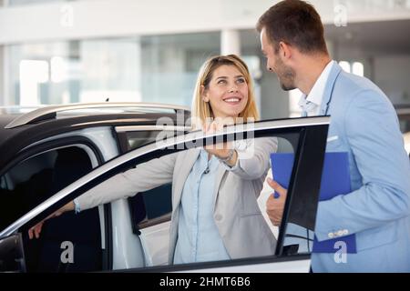 Giovane donna che acquista una nuova auto in concessionaria città e parla con il venditore Foto Stock