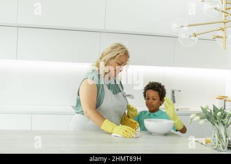 La famiglia felice pulisce la camera. Madre e figlio fanno la pulizia in casa. Donna matura e un ragazzino che si divertono e puliscono la cucina Foto Stock