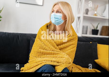 Donna anziana malata che indossa maschera protettiva viso sensazione di malessere a casa, si siede sul divano coperto di coperta, si sente brividi e febbre, una femmina ha malattia e auto isolato a casa Foto Stock