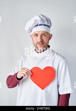 Cuoco o fornaio maschile allegro in uniforme bianca con cuore di carta rosso. Ristorante di concetto caffè. Cuoco chef che tiene un cuore rosso di fronte all'abbigliamento da lavoro. Foto di alta qualità Foto Stock