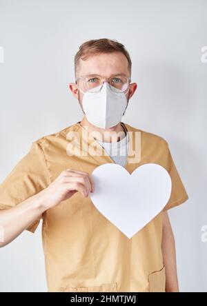 Ritratto di un medico cardiologo maschile o infermiere in maschera medica con carta bianca forma del cuore. Medico caucasico professionista in ospedale. Medico chirurgo adulto in ospedale. Foto verticale di alta qualità Foto Stock