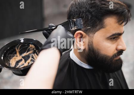 Barbiere professionale che moriva i capelli del cliente maschile in barbiere. Il parrucchiere applica la tintura ai capelli del cliente con una spazzola Foto Stock