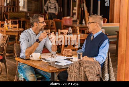 Due uomini d'affari senior sorridono mentre parlano al bar e bevono il loro caffè Foto Stock