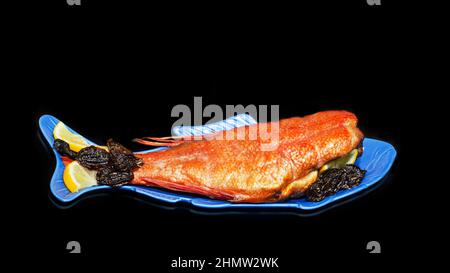 Spigola affumicata calda con limone e prugne secche, primo piano su un piatto a forma di pesce e sfondo nero. Frutti di mare deliziosi per una corretta alimentazione Foto Stock