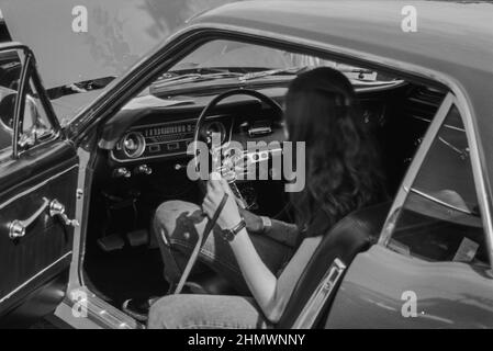 Foto in scala di grigi di una giovane donna sulla vecchia auto classica Ford Mustang in una strada Foto Stock